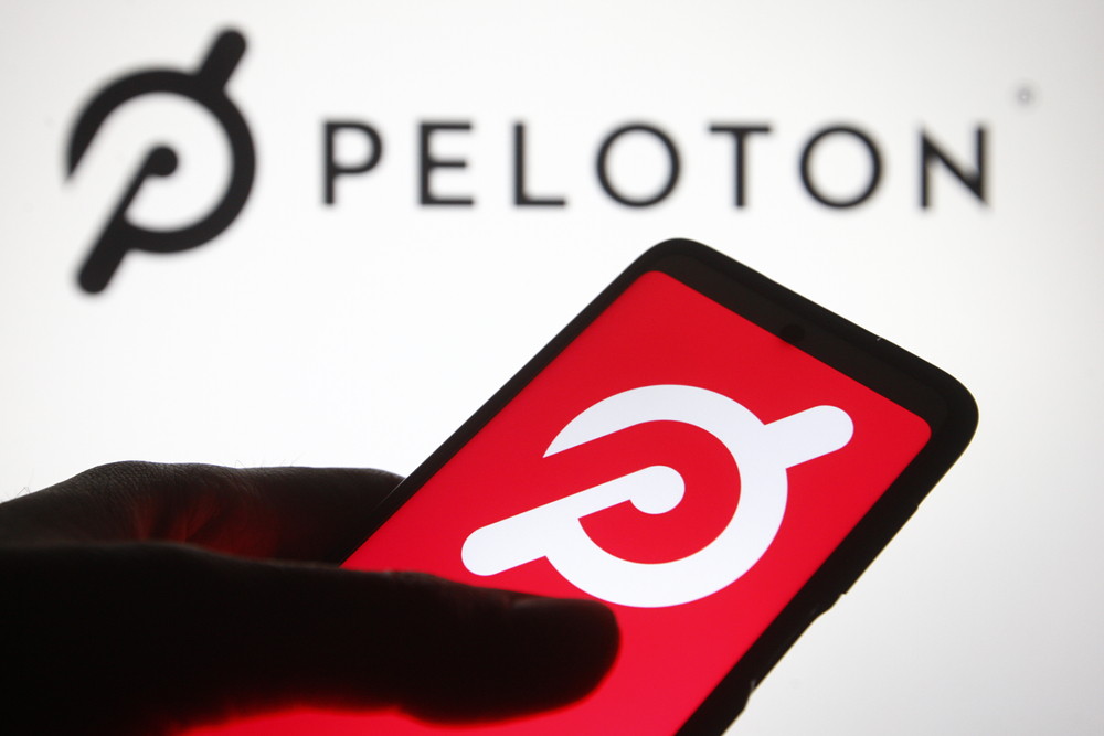 a hand holding a phone with Peloton logo underneath Pelton logo on wall