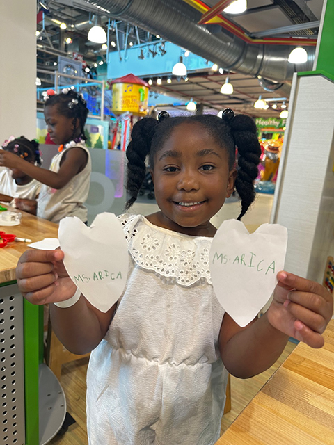 litle girl smiling and holding homemade paper hearts reading 'Ms. Arica