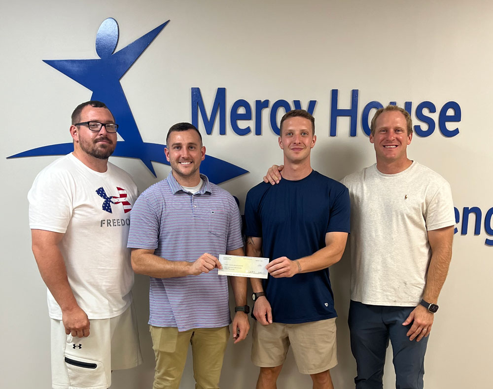 four men standing in front of a Mercy House sign, two holding a donation check
